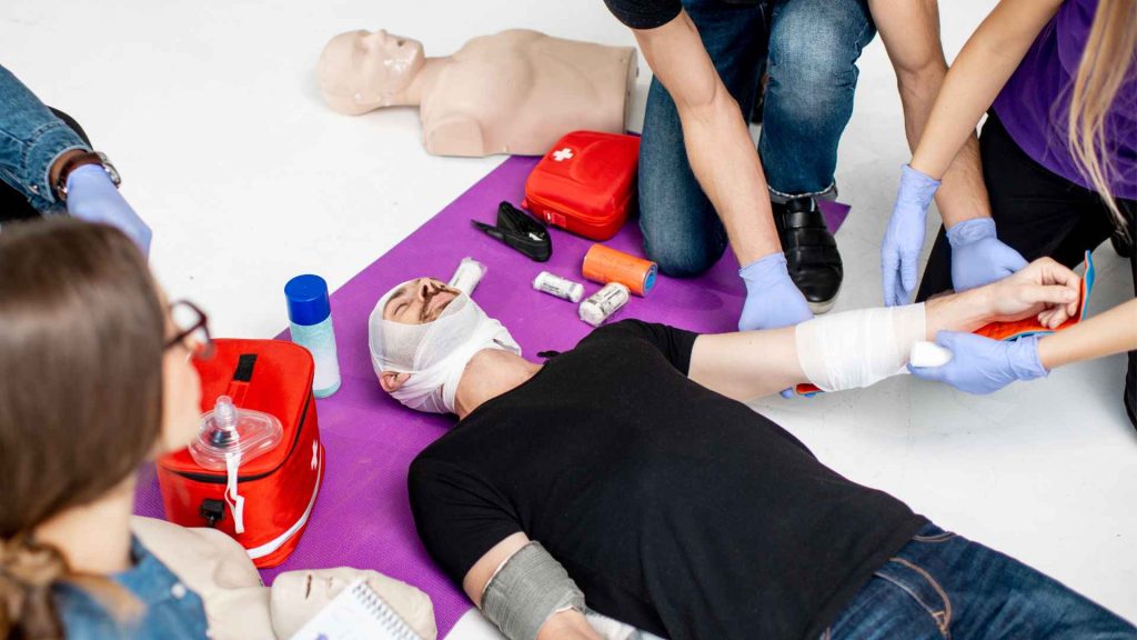 students in a first aid training class practicing their bandaging skills. the students are practicing on one man who is laying on the floor. his head is wrapped in bandages and someone is currently bandaging his arm.