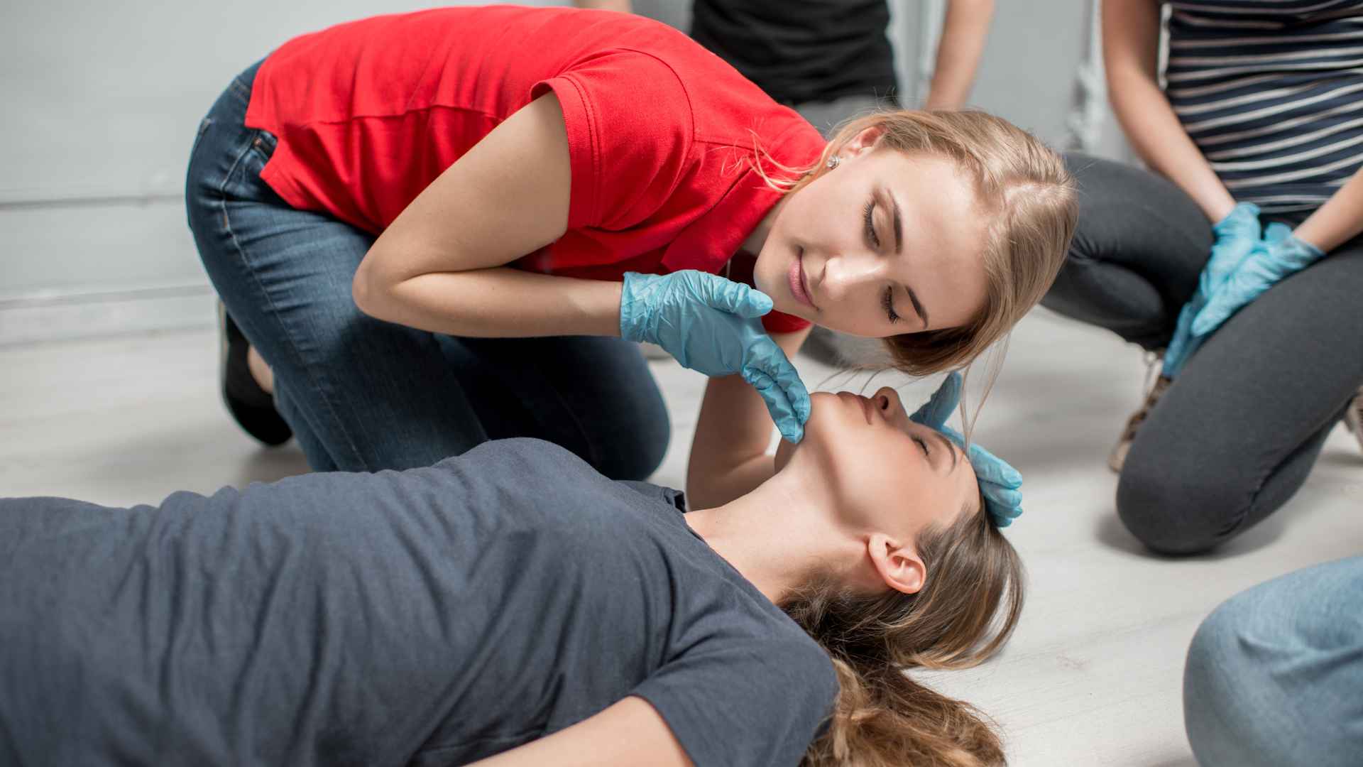 woman in red shirt wearing blue nitrile gloves performing a head-tilt-chin-lift on another woman laying on the ground. the woman in red is listening for the other woman's breathing and checking to see if her chest is rising and falling as part of CPR's ABCs.