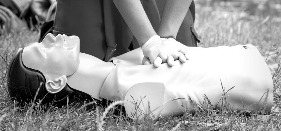 Person practicing CPR on a training mannequin outdoors. The individual's gloved hands are positioned on the mannequin's chest, simulating chest compressions. The scene is set in a grassy area.