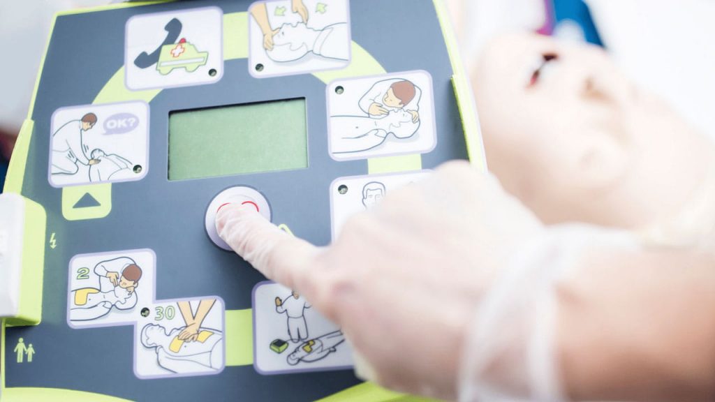 A hand wearing a glove presses a button on a defibrillator, which displays illustrated instructions for use. A mannequin head is partially visible in the background, indicating a training scenario.