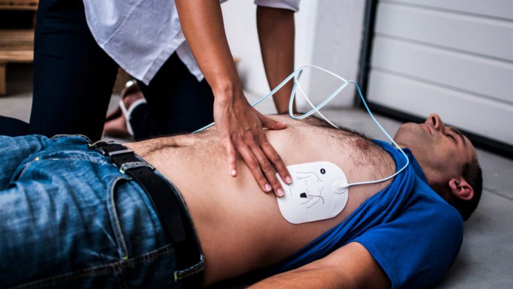 A person performs CPR on a man lying on the ground. The man has a defibrillator pad on his chest, wearing a blue shirt and jeans. The other person's hands are pressing on his chest for resuscitation.