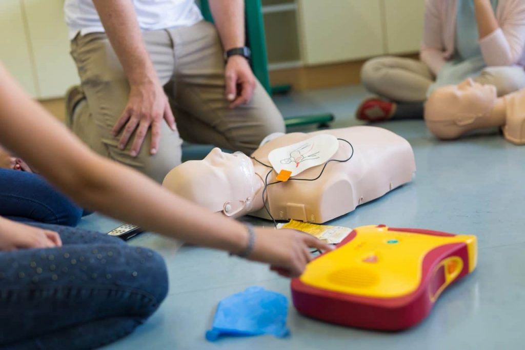 People are gathered around a CPR training mannequin and a defibrillator. One person points at the defibrillator, while others observe. The setting appears to be a training session, with the focus on emergency response techniques.