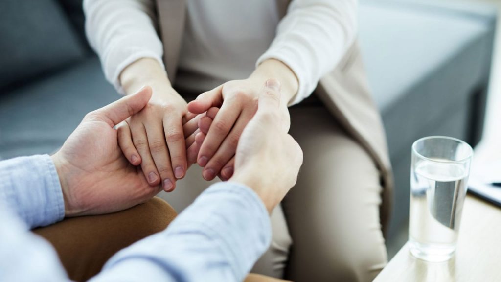 Close-up of two people sitting on a couch, holding hands in a comforting manner. One person gently wraps their hands around the other's. A glass of water is on a table nearby. Their attire is casual.