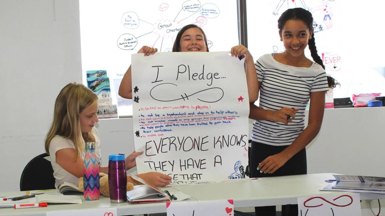 Three girls in a classroom setting, one seated with a notebook, and two standing, holding a large poster that reads "I Pledge... Everyone knows they have a voice..." The background shows a window and a whiteboard with doodles.