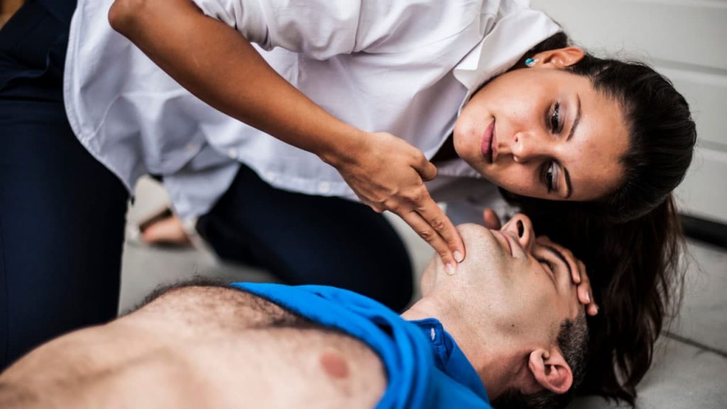 A woman in a white shirt checks the breathing of an unconscious man lying on the ground. She tilts his head back and holds his chin while leaning over him. The man is wearing a blue shirt.
