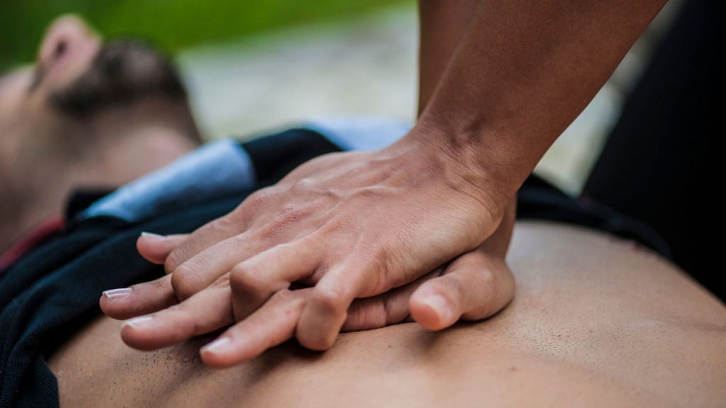Close-up of a person's hands performing CPR chest compressions on another person's chest. The person receiving CPR is lying on their back, and the image focuses on the hands and upper body, conveying a sense of urgency and care.