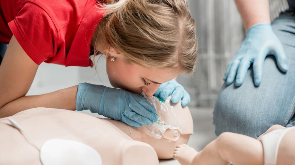 A person wearing a red shirt and blue gloves practices giving rescue breaths on a CPR mannequin. Another person in jeans and gloves is visible nearby, observing or assisting with the training session.