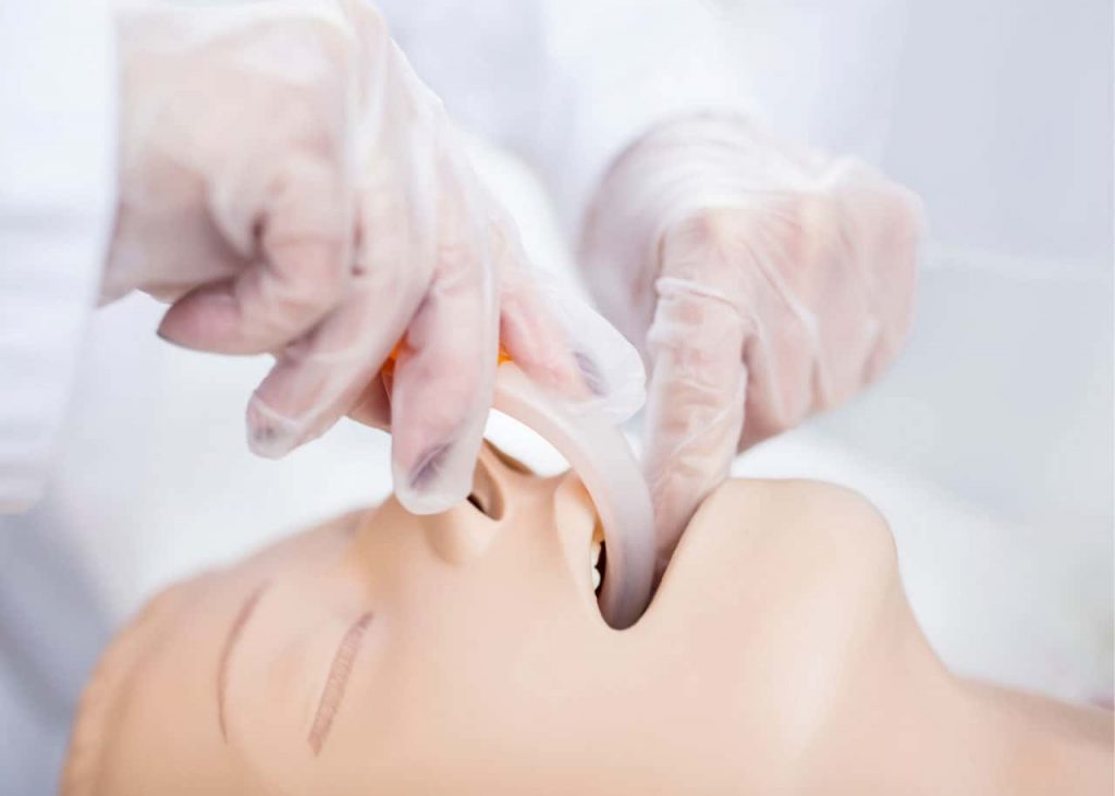 A person wearing gloves inserts a medical airway device into a mannequin's mouth, demonstrating an intubation procedure. The mannequin is shown from a side angle focusing on the face and mouth.