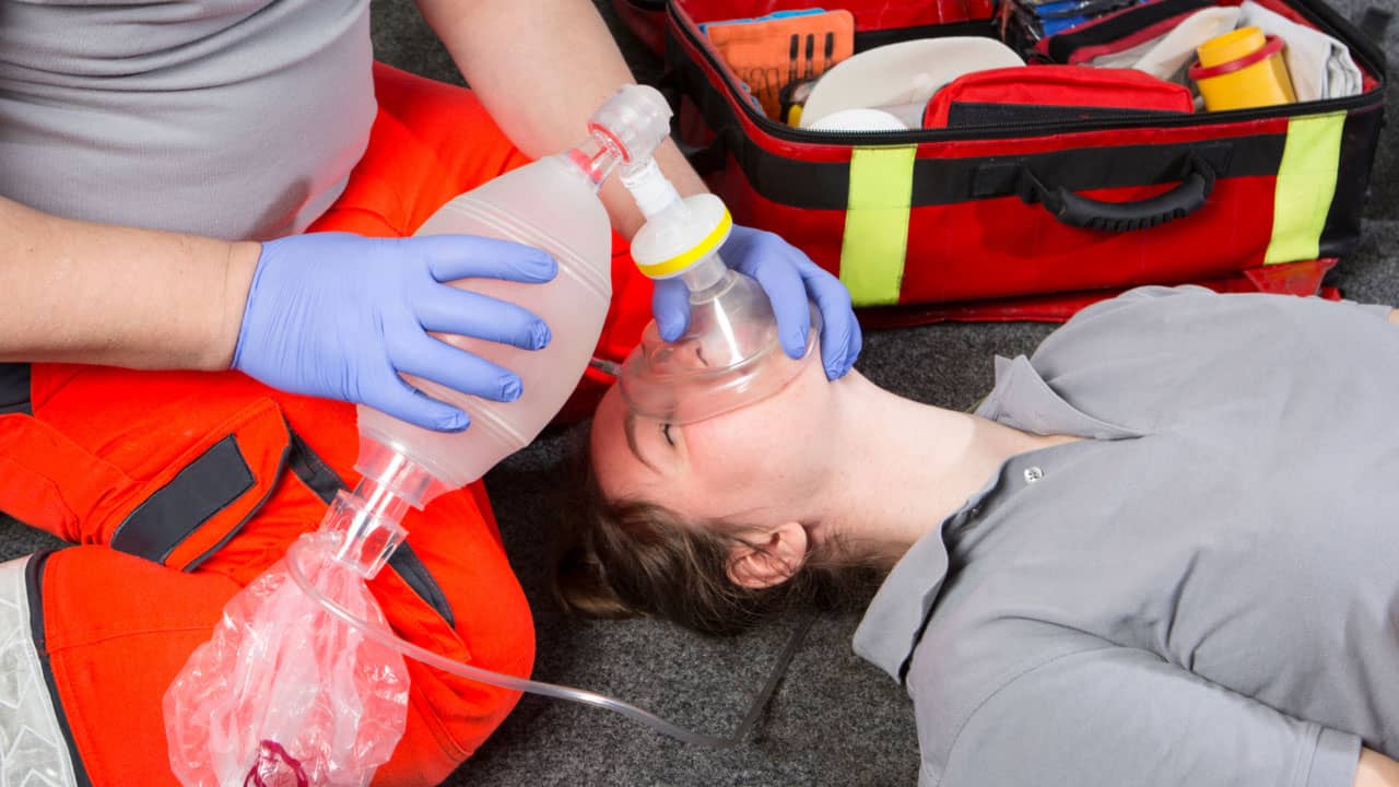 A person in a gray shirt receiving CPR, with another person wearing orange pants and blue gloves using a bag valve mask to administer breaths. A medical kit is open on the ground nearby.