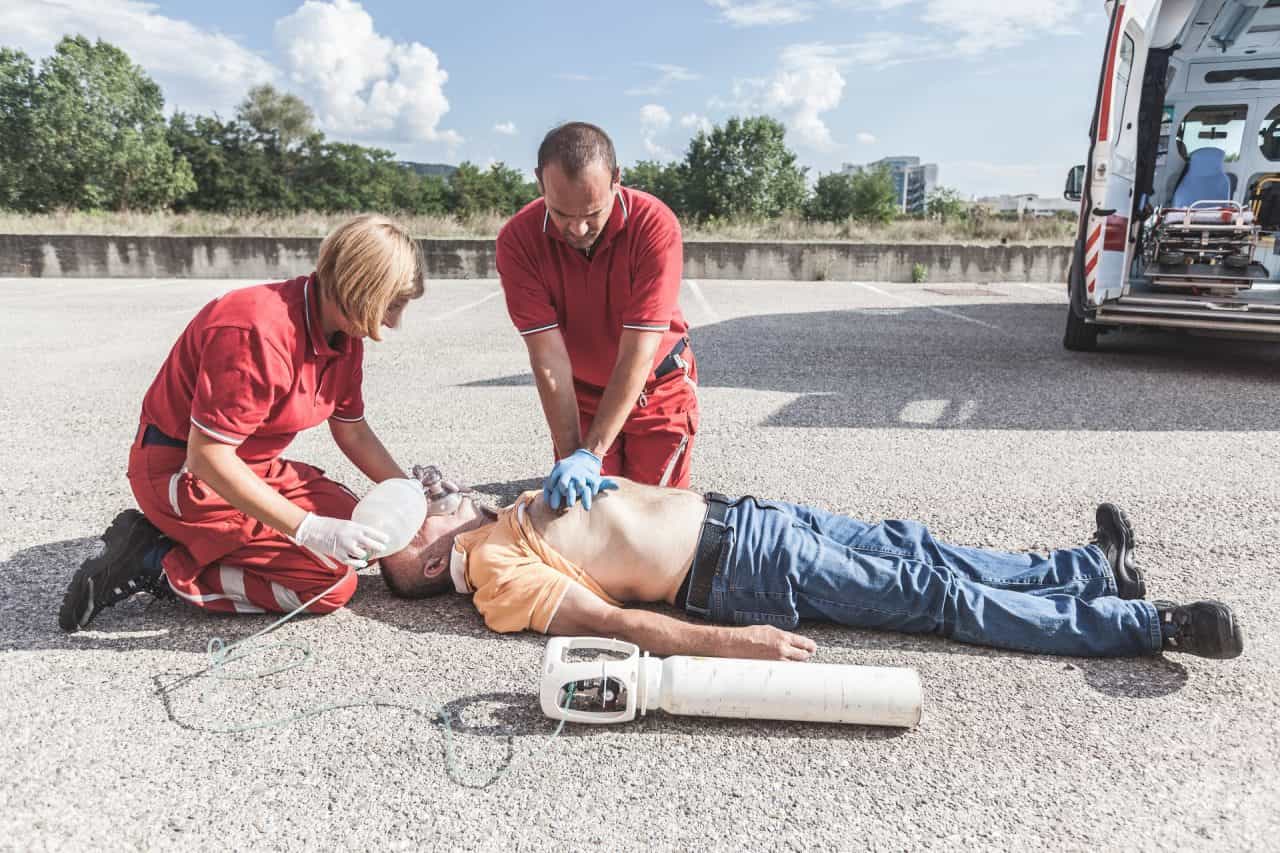 Two emergency responders in red uniforms perform CPR on a man lying on the ground. One administers chest compressions while the other uses a bag valve mask for ventilation. An ambulance is partially visible nearby.