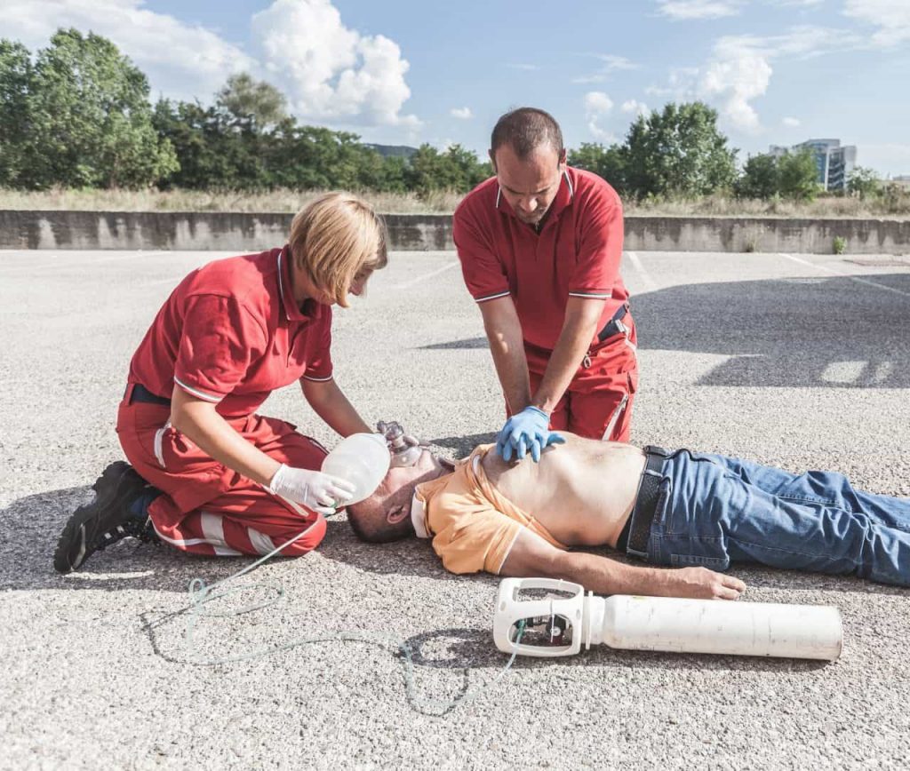 Two rescuers in red uniforms perform CPR on a man lying on the ground. The man is shirtless with an oxygen mask placed by one rescuer, while the other performs chest compressions. An oxygen tank is beside them on the pavement.