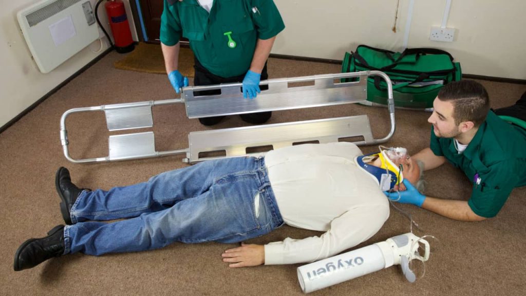 Two paramedics are assisting a person lying on the floor with a neck brace and oxygen mask. One paramedic is holding a stretcher, while the other is adjusting the mask. An oxygen tank is nearby.
