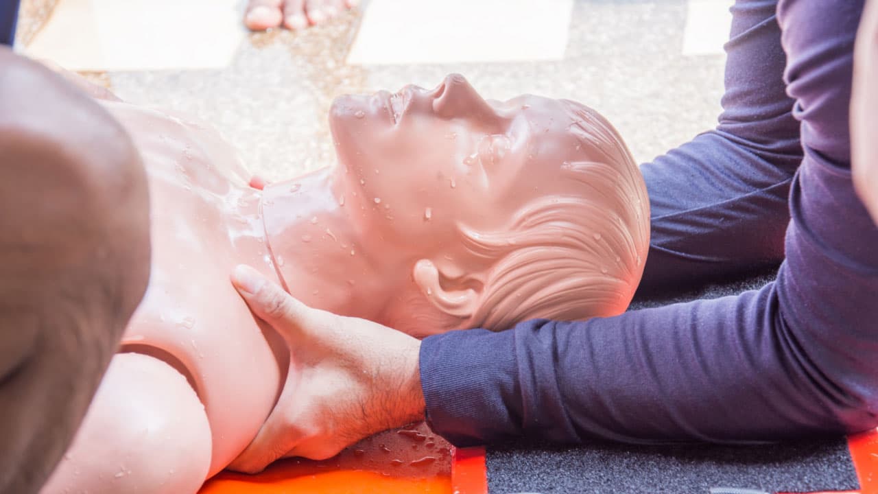CPR training on a dummy, with two people's hands positioning the head for airway management. The dummy is on a red board, and the scene is well-lit, focusing on the importance of life-saving techniques.
