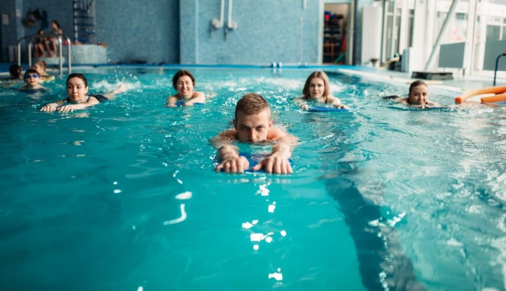 A group of people swimming in a pool, using kickboards. They are aligned in a row, each focusing on swimming. The pool area has blue tiles and a modern look, with some equipment visible in the background.