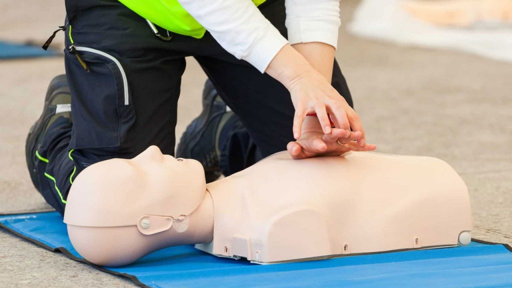 A person in a reflective vest kneels on a mat practicing CPR on a training mannequin by pressing down on its chest with interlocked hands.