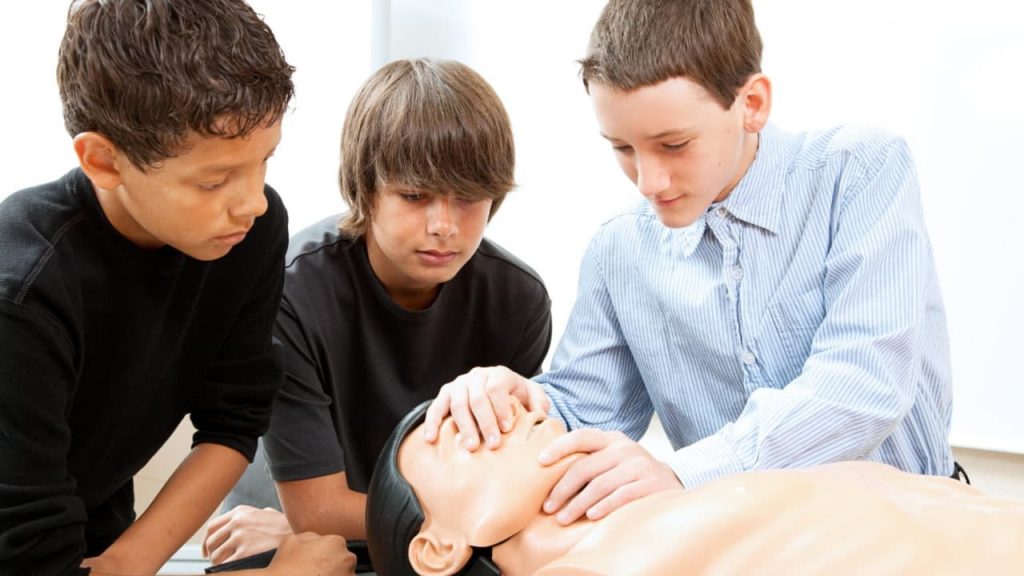 Three boys are practicing CPR on a training dummy. One boy holds the dummy's nose and chin, while another observes closely. They are focused on learning the procedure in a classroom setting.