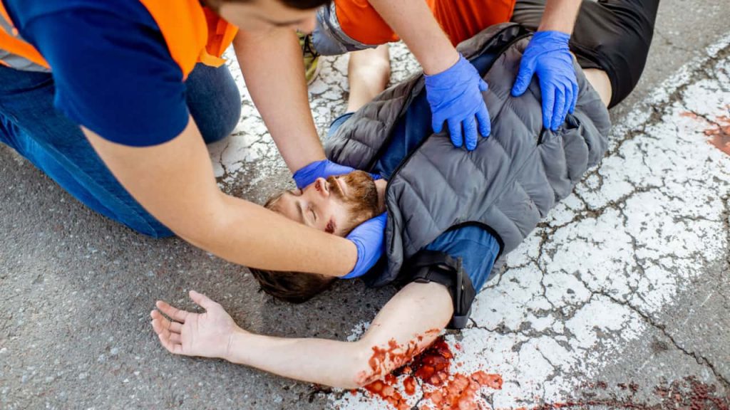 Two individuals in orange safety vests and blue gloves are assisting an injured person lying on a cracked, blood-stained pavement. One holds the person's head, while the other checks their torso. The person’s arm is outstretched.