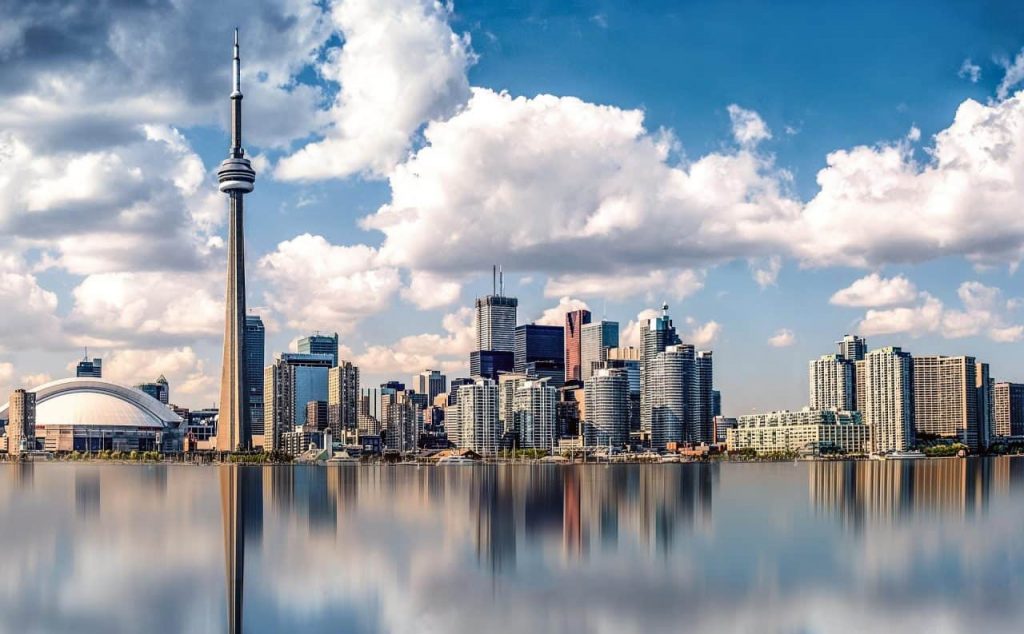Toronto skyline with the CN Tower and various skyscrapers under a blue sky with clouds. Buildings are reflected in the calm water in the foreground, creating a mirrored effect.