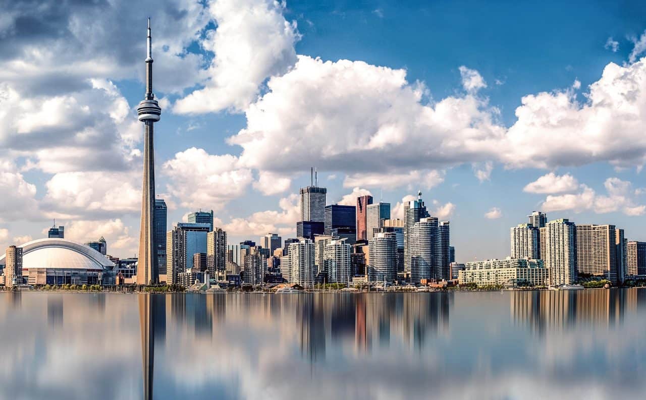 Toronto skyline with the CN Tower and various skyscrapers under a blue sky with clouds. Buildings are reflected in the calm water in the foreground, creating a mirrored effect.