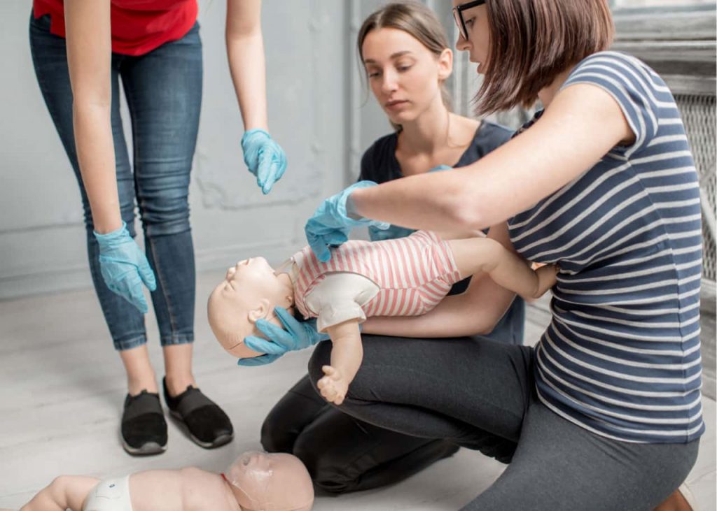 Three people wearing gloves practice infant CPR on a baby mannequin. One person holds the mannequin, while another assists. A second mannequin lies on the floor nearby. They appear focused and attentive.