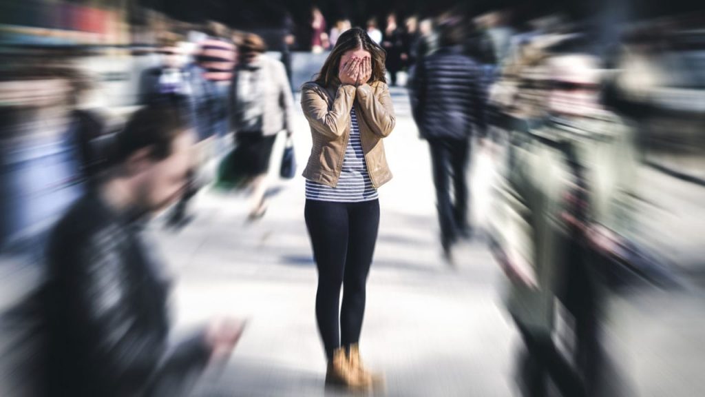 A person stands overwhelmed in a busy street, covering their face with their hands. The background is blurred, highlighting the sense of anxiety amidst the motion of people passing by.