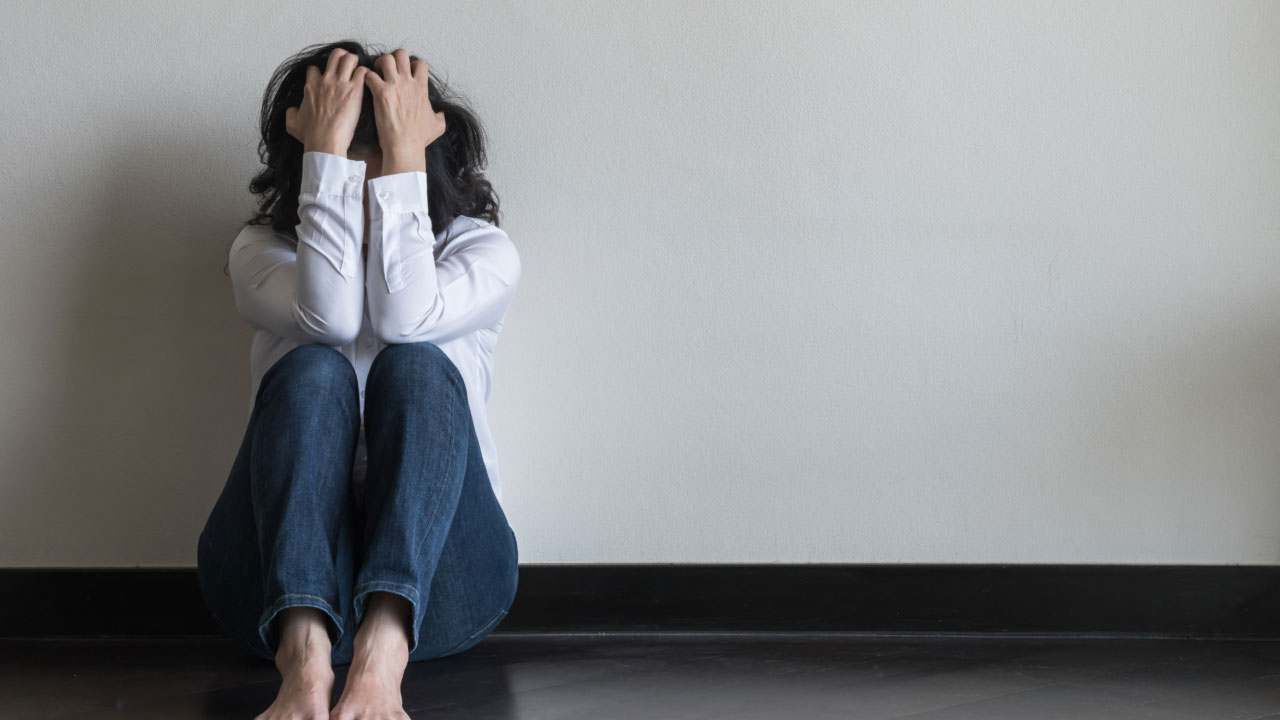 A person sits on the floor against a plain wall, dressed in a white shirt and blue jeans. They have their knees up and head in hands, conveying a sense of distress or overwhelming emotion. Barefoot, they appear deep in thought or anxiety.
