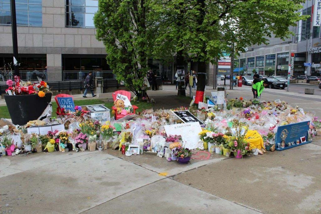 A large memorial on a city sidewalk features numerous flowers, candles, stuffed toys, and heartfelt messages. People walk nearby, and buildings are visible in the background.