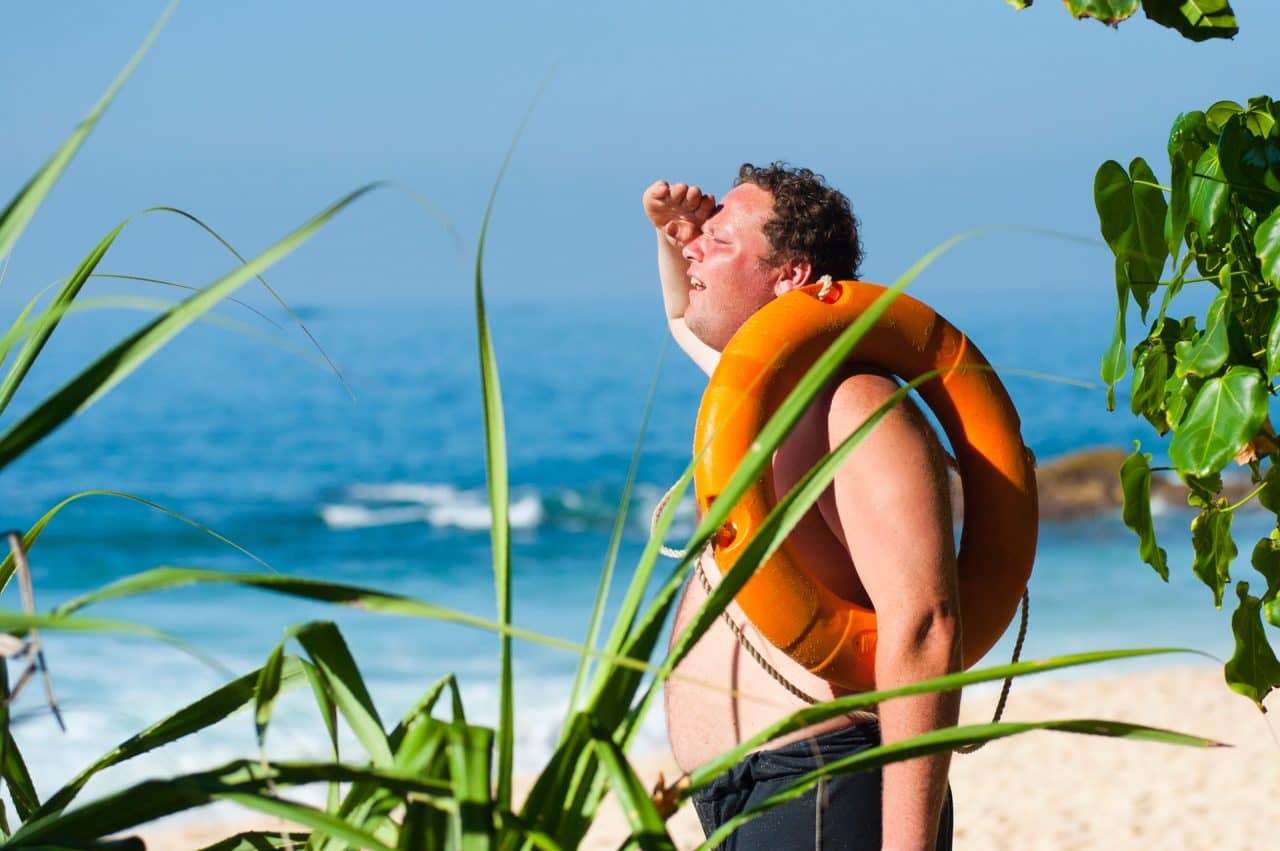 A man wearing an orange lifebuoy stands on a sandy beach, shielding his eyes from the sun. He faces the ocean, surrounded by greenery. The sea is calm, and the sky is clear and bright.