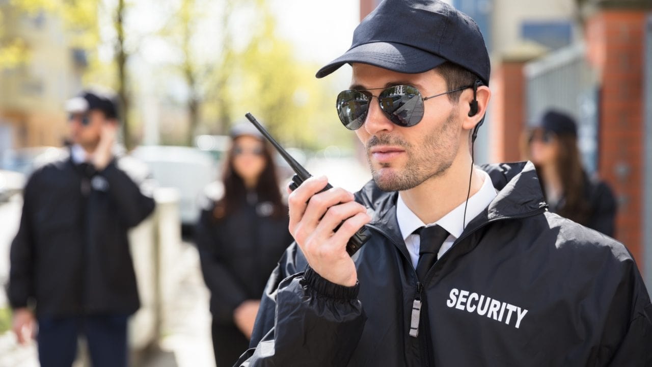 A man wearing sunglasses and a cap, dressed in a security jacket, talks into a walkie-talkie. In the background, three other security personnel stand out of focus. They all appear to be outdoors, near a building.