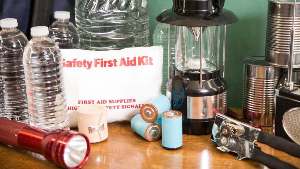 Emergency preparedness supplies including bottled water, a "Safety First Aid Kit," batteries, a red flashlight, a lantern, canned goods, a can opener, and a radio are arranged on a table.
