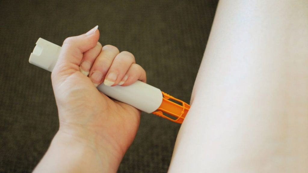 A person holds an auto-injector pen against their leg, preparing to administer a shot. The pen is white with an orange tip. The background is a neutral carpeted floor.