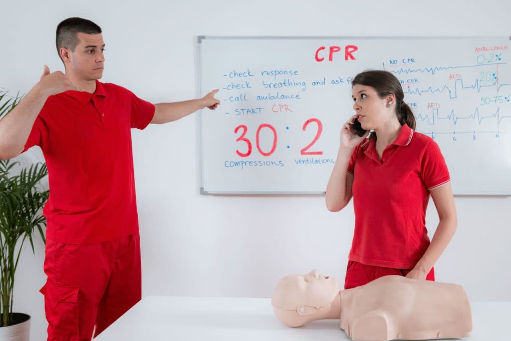 A man in red demonstrates a hand signal, standing near a woman on the phone, in front of a CPR instruction board. A CPR dummy is on the table. The board lists CPR steps and the compression-to-breath ratio, "30:2.