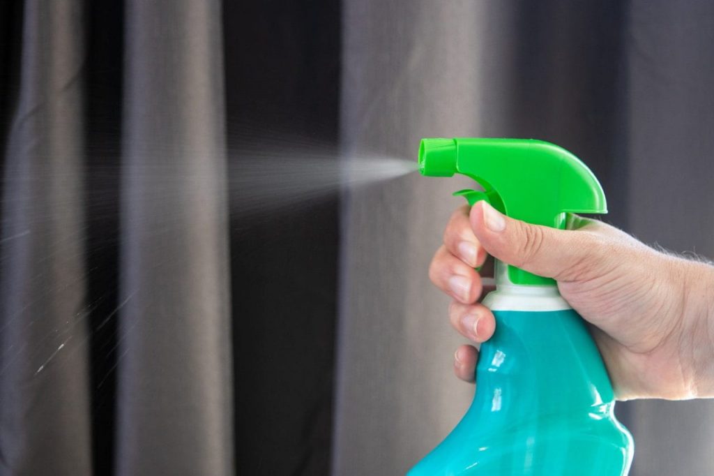 A hand is holding a green spray bottle, releasing a mist of liquid. The background consists of dark, blurred curtains.