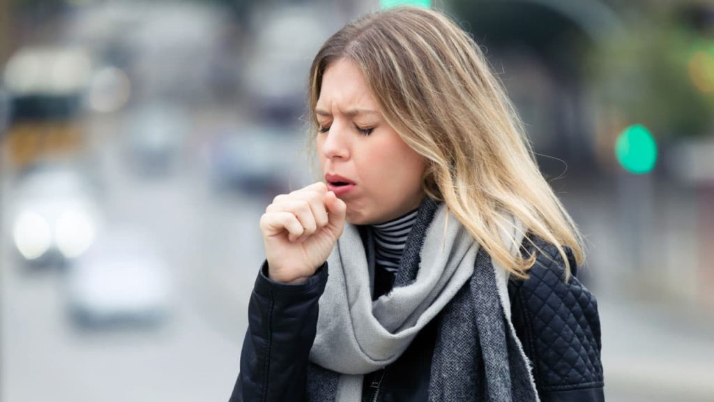 A woman with long blonde hair is standing outdoors, coughing into a closed fist. She is wearing a black jacket, a gray scarf, and a striped turtleneck. The background is a blurred city street with traffic lights.