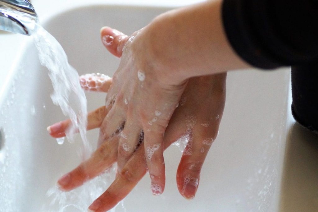 A person washing hands with soap under running water in a white sink, creating bubbles and foam. The image focuses on the hands and the flow of water, emphasizing cleanliness and hygiene.