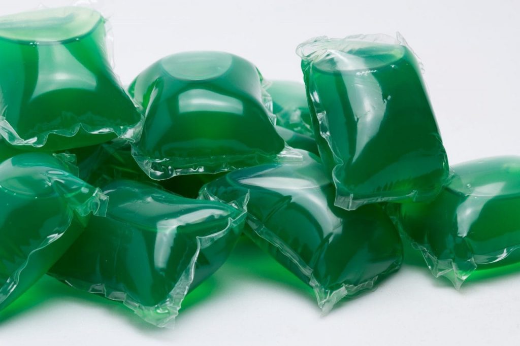 A pile of green liquid-filled detergent pods, each wrapped in a transparent film, are scattered on a white surface.