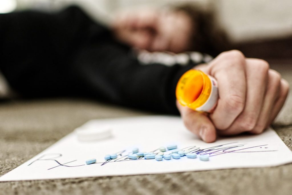 A person lying on the floor, holding an open prescription bottle in one hand. Pills are scattered on a paper with a prescription symbol. The focus is on the pills and the bottle, with the person's face blurred in the background.