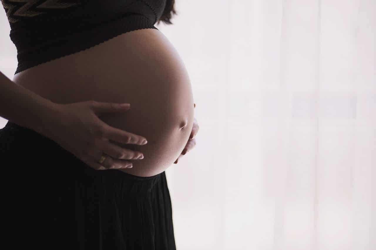 A close-up side view of a pregnant person gently holding their bare belly with both hands. They are wearing a black top and skirt. Soft light filters through a sheer curtain in the background.