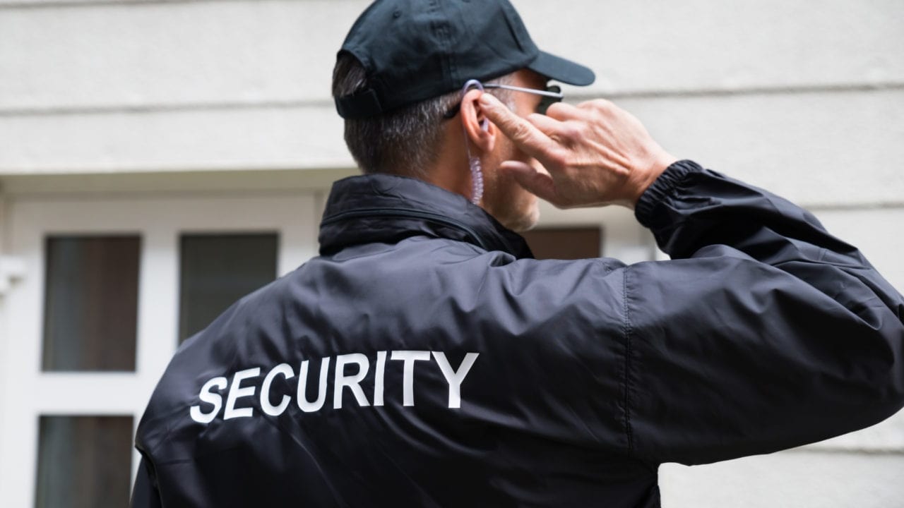 A security guard wearing a black jacket with "SECURITY" written on the back and a black cap stands outside a building. They are holding an earpiece with their right hand, appearing attentive and focused.