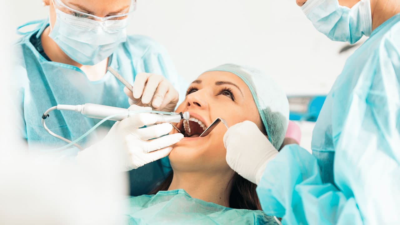 A woman is receiving dental treatment from two dentists. She is lying back with her mouth open as they work, wearing blue surgical gowns, gloves, and masks. The setting appears to be a dental clinic.