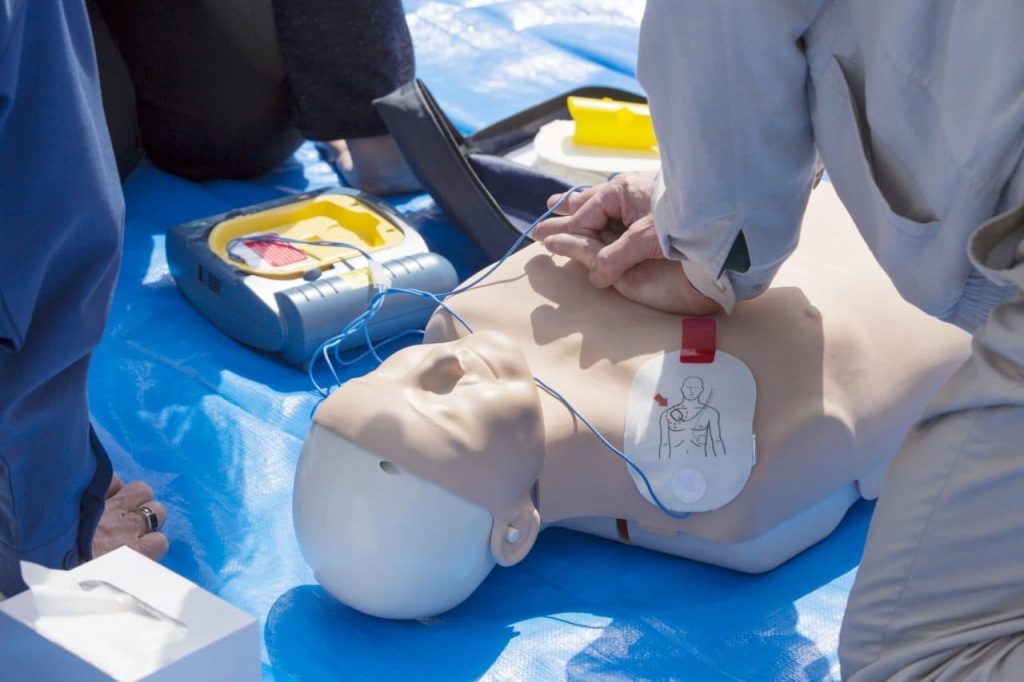 Person demonstrating CPR on a mannequin with an AED device next to them, placed on a blue tarp. The mannequin has electrode pads on its chest, and the scene indicates a training session.