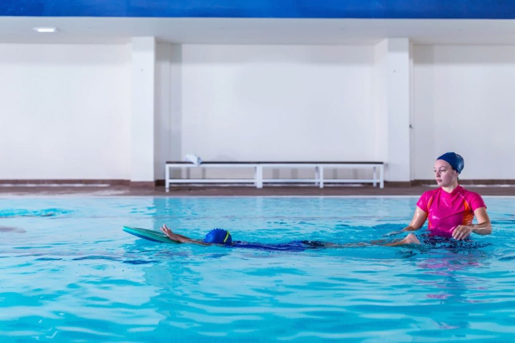 A swimming instructor in a pink shirt guides a child in a blue swimsuit using a kickboard in an indoor pool. The child is floating on their stomach, and the instructor is providing support while standing in the water.
