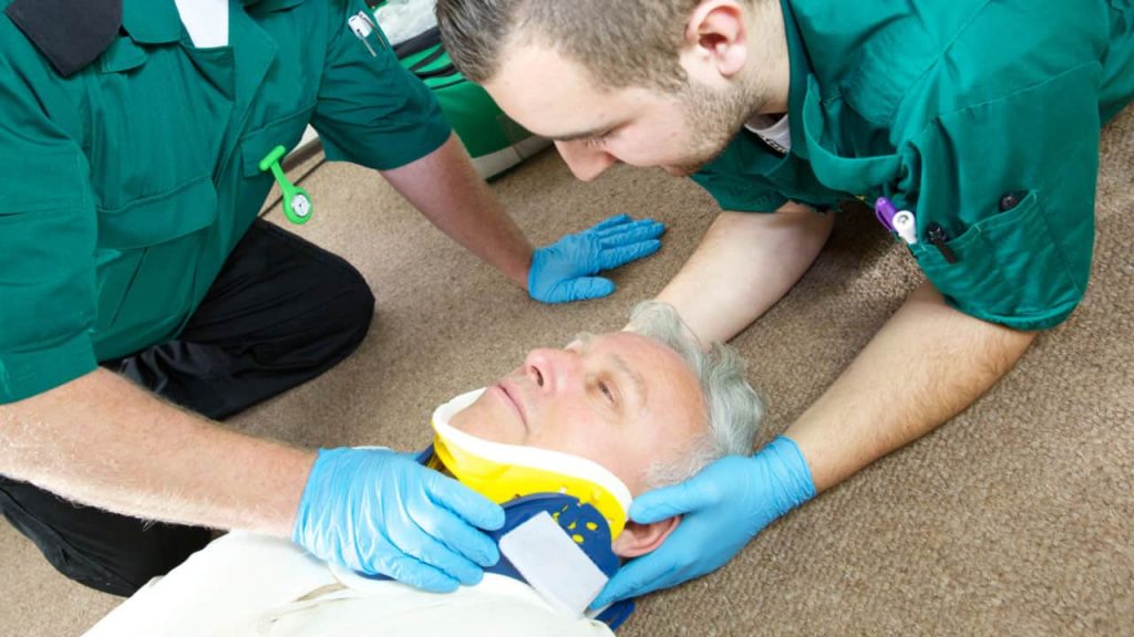Two paramedics assist an older man lying on a carpeted floor. They are securing a yellow and blue neck brace on him. Both paramedics wear green uniforms and blue gloves, focusing intently on providing care.