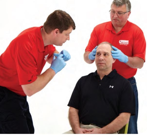 Two men wearing red shirts and blue gloves examine the head of a seated man in a black shirt. One of them is leaning forward, closely inspecting, while the other holds the seated man's head steady.