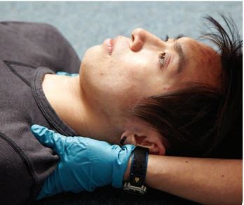 A person lying on their back on a blue surface, wearing a dark shirt. They have a blue gloved hand gently supporting the back of their head, possibly in a medical or caring context.