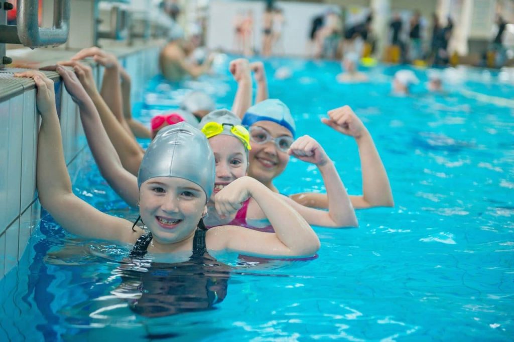 Four children wearing swim caps and goggles stand in a row at the edge of a swimming pool. They smile and flex their arms in a playful manner. The pool is busy with other swimmers in the background.
