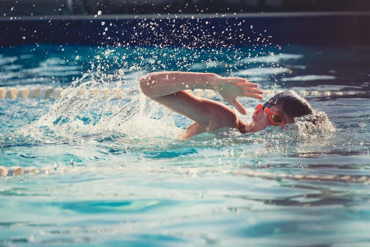 A person wearing goggles swims freestyle in an outdoor pool, creating splashes of water. The sunlight reflects off the water surface, and lane dividers are visible in the background.