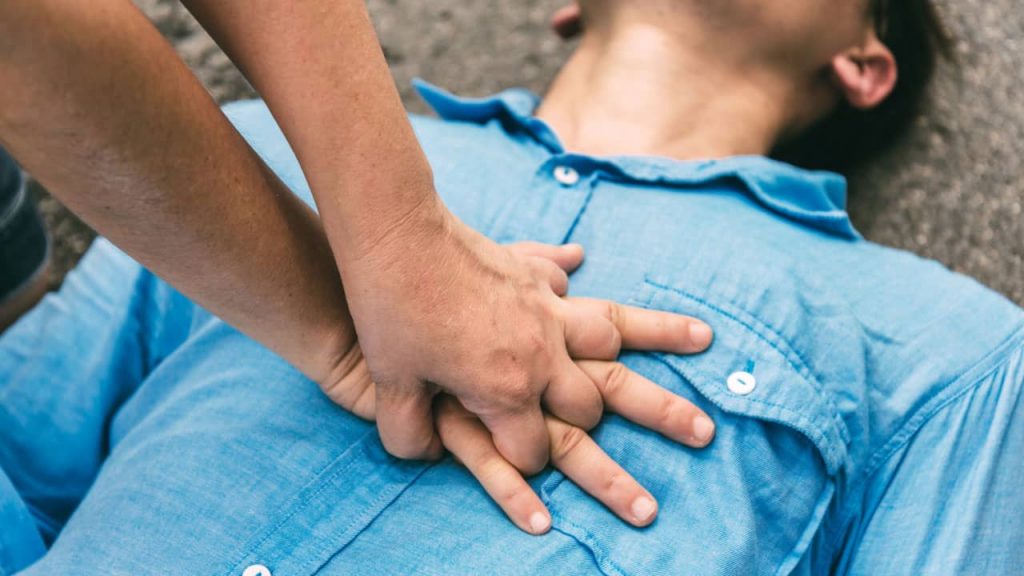 A person in a blue shirt is receiving CPR on a gray surface. Hands are pressing on their chest, demonstrating a lifesaving technique. The scene suggests urgency, focusing on the chest compression process.