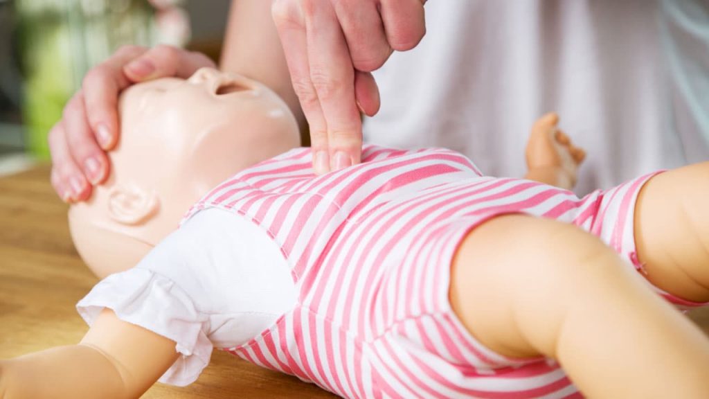 A person demonstrates CPR on an infant mannequin. The mannequin is dressed in a pink and white striped outfit. One hand supports the mannequin's head, while two fingers perform chest compressions.
