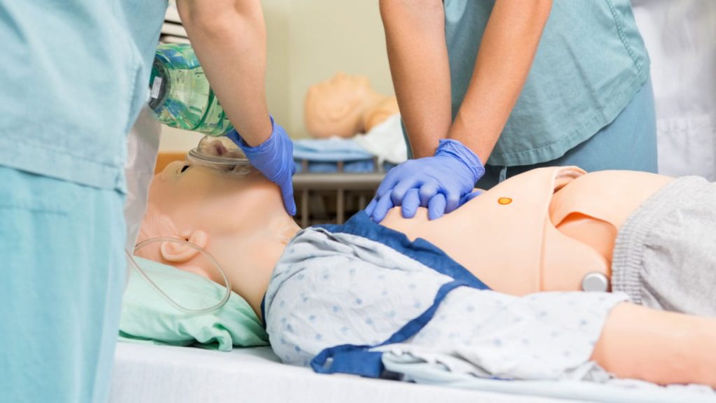 Medical professionals in scrubs and gloves practice CPR on a medical training mannequin, focusing on chest compressions and ventilation. Additional medical equipment is visible in the background.